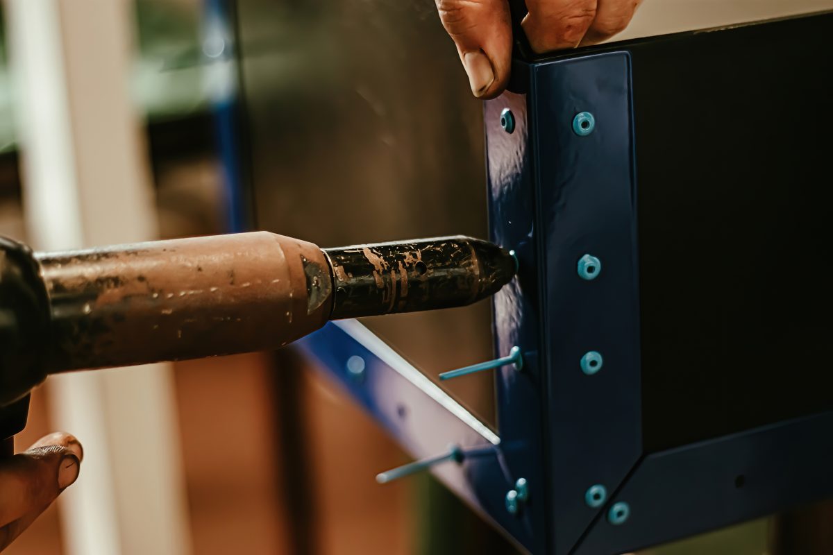 Proces nitowania z wykorzystaniem nitownicy, widoczna ręka operatora i metalowy panel z zamontowanymi nitami