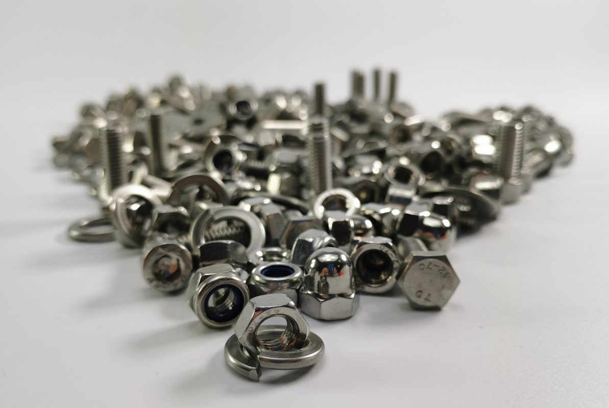 Zbliżenie na różne elementy złączne, takie jak nakrętki, śruby i podkładki, wykonane ze stali nierdzewnej. Ilustracja porównawcza dotycząca właściwości i różnic między stalą A2 a stalą A4, używaną w przemyśle do produkcji wytrzymałych elementów odpornych na korozję.